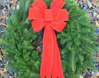 May include: A circular evergreen wreath with a vibrant red bow. The wreath is made of fresh, green pine needles and is adorned with a large, plush red bow. The bow's long tails cascade down, adding a festive touch. Perfect for holiday decor.