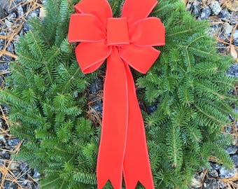 May include: A fresh evergreen wreath with a vibrant red bow. The wreath is made of lush green pine needles and features a large, decorative bow with long, flowing tails. Perfect for holiday decor.