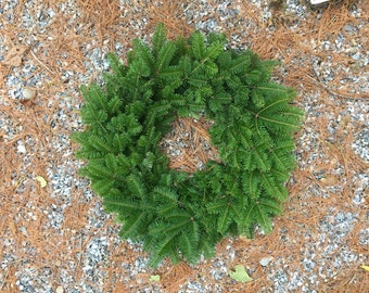 May include: A fresh, circular evergreen wreath composed of vibrant green fir branches. The wreath is displayed on a bed of small gray stones and scattered brown pine needles, creating a natural, seasonal aesthetic.