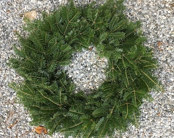 May include: A fresh, circular evergreen wreath composed of lush green fir branches. The wreath is displayed on a bed of small, gray pebbles, creating a natural and rustic aesthetic. Ideal for seasonal home decor.
