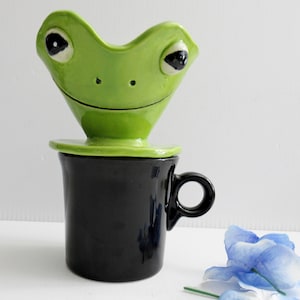 May include: A ceramic coffee pour-over dripper shaped like a green frog head with black eyes and a smiling mouth, resting securely on top of a solid black cylindrical coffee mug with a circular handle, featuring a glossy glazed finish.