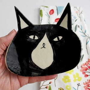 May include: A black and white ceramic cat dish with a white face and black ears. The dish is shaped like a cat's head and has a glossy finish.