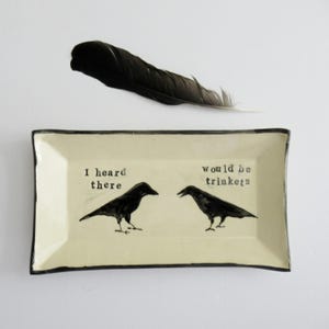 May include: A rectangular off-white ceramic tray with a dark rim featuring two black crow illustrations and the text "I heard there would be trinkets" printed in black. A single black feather is positioned above the tray.