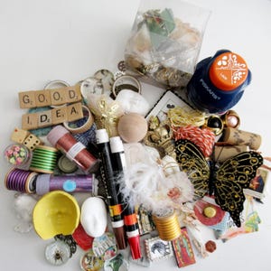 May include: A collection of craft supplies including wooden Scrabble tiles spelling GOOD IDEA, a Fiskars lace punch, a large black and gold butterfly ornament, two markers, spools of thread, glitter tubes, small white face masks, and various decorative trinkets.