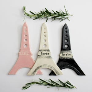 May include: Three Eiffel Tower-shaped ceramic herb markers in pink, cream, and black. Each marker has the word "herbe" and decorative trim. Rosemary sprigs are placed above and below the markers.