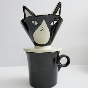 May include: A black and white ceramic coffee pour-over dripper shaped like a cat head, resting on a solid black cylindrical mug with a circular handle. The cat-shaped dripper features pointed ears, almond-shaped eyes, and a white muzzle area.