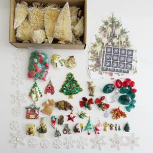 May include: A collection of Christmas-themed craft supplies including a box of miniature bottle brush trees, a bag of assorted green and red buttons, various holiday-themed charms, resin figurines, faceted plastic gems, and white snowflake cutouts.