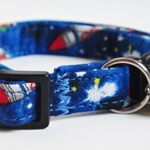 May include: Blue cat collar with a space-themed print featuring rockets and stars. The collar has a black buckle and a silver bell.