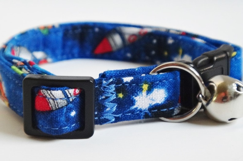 May include: Blue cat collar with a space-themed print featuring rockets and stars. The collar has a black buckle and a silver bell.