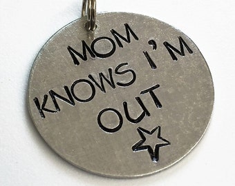 May include: Silver round charm with a black inscription that reads "Mom Knows I'm Out" with a star below the text.