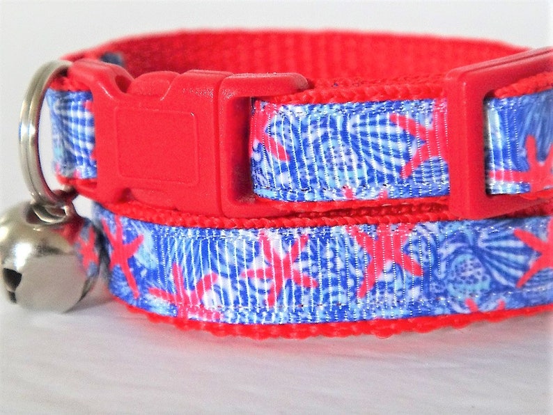 Peut inclure: Collier de chat rouge avec un ruban &agrave; motif bleu et blanc orn&eacute; d'&eacute;toiles de mer roses. Le collier a une boucle rouge et une clochette argent&eacute;e.