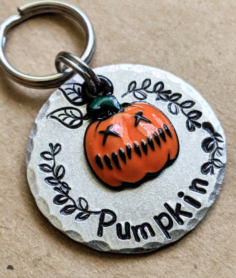 May include: A silver metal pet tag with a black and orange pumpkin design and the text "Pumpkin" in a decorative font.