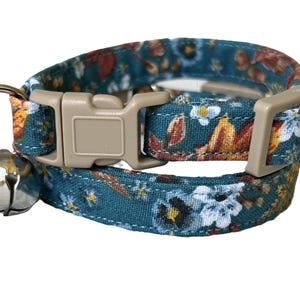 May include: A teal blue cat collar with a floral pattern featuring white, orange, and brown flowers. The collar has a beige buckle and a silver bell.