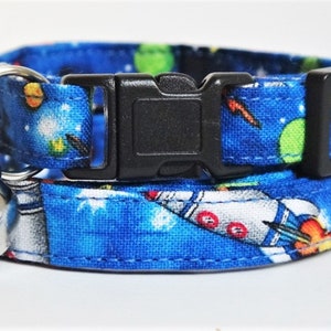 May include: A blue cat collar with a space-themed print featuring rockets, planets, and stars. The collar has a black buckle and a silver bell.