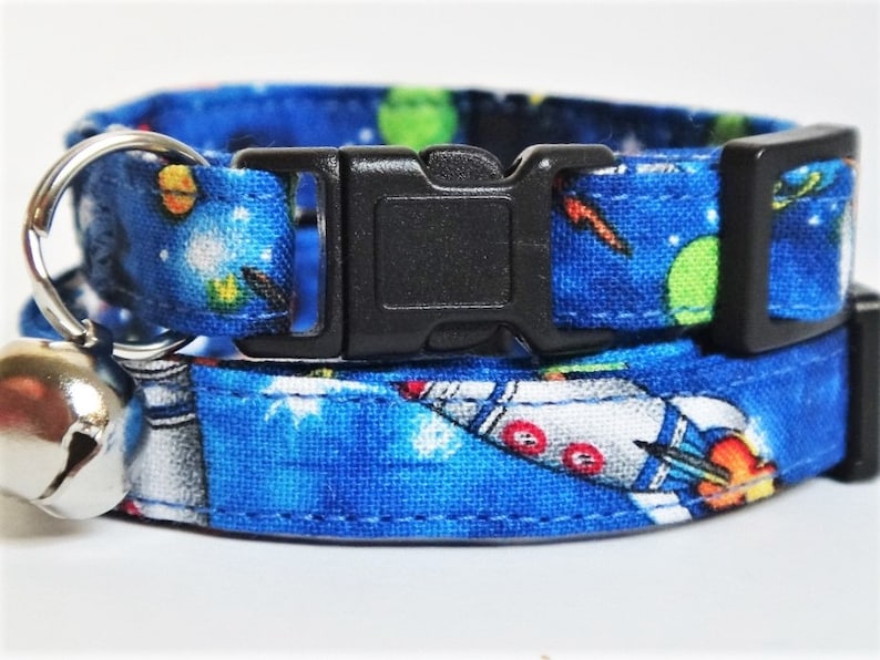 May include: A blue cat collar with a space-themed print featuring rockets, planets, and stars. The collar has a black buckle and a silver bell.