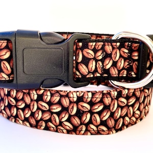 May include: A dog collar featuring a pattern of coffee beans in shades of brown and tan against a black background. The collar has a black plastic buckle and a silver metal D-ring.