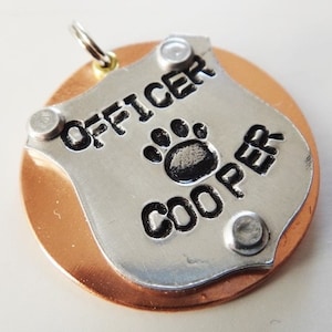 Puede incluir: Chapa de perro de plata con el texto "OFFICER COOPER" y una huella de pata. La chapa est&aacute; unida a una base redonda de color cobre.