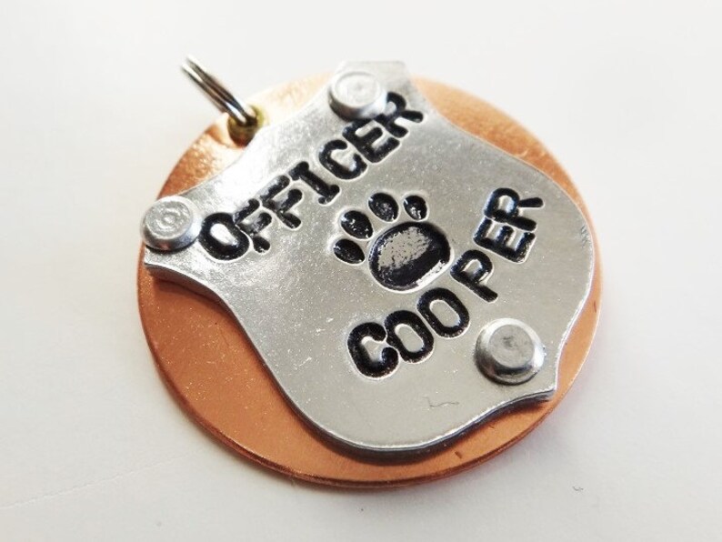 Puede incluir: Una placa de perro de plata con las palabras "OFFICER COOPER" y una huella de pata. La placa est&aacute; unida a un disco de color cobre.