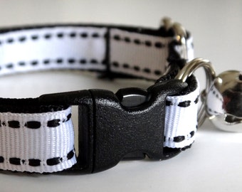 May include: White and black cat collar with a black buckle and a silver bell. The collar is made of a soft, comfortable material and has a safety release buckle.