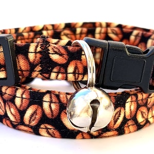 May include: A brown and black cat collar with a coffee bean pattern and a silver bell. The collar has a black plastic buckle.