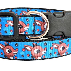 May include: A blue dog collar with a festive reindeer pattern and red and black polka dots. The collar has a black plastic buckle and a silver metal D-ring.
