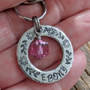 May include: Silver key chain with a pink crystal charm and the word "EBONY" engraved on a circular tag. The tag is decorated with a floral design.