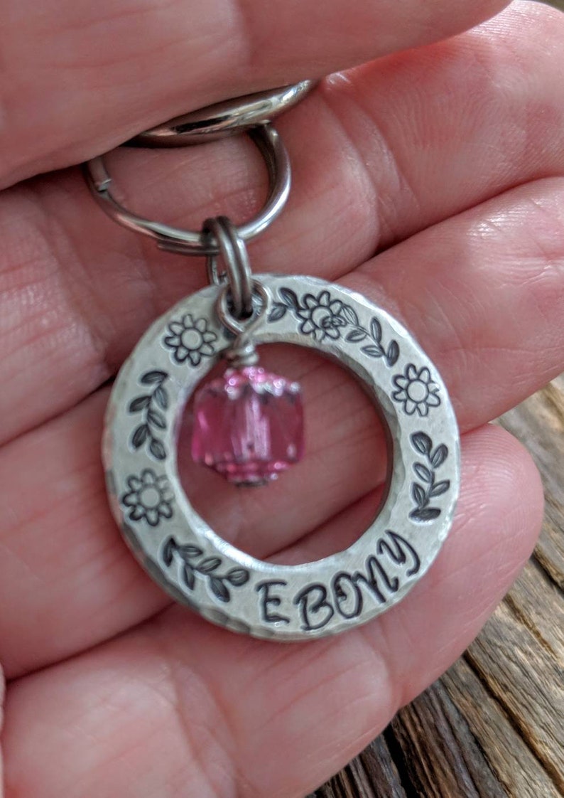 May include: Silver key chain with a pink crystal charm and the word "EBONY" engraved on a circular tag. The tag is decorated with a floral design.