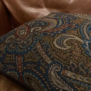 Puede incluir: Una almohada de tiro de color marr&oacute;n, azul y dorado con un patr&oacute;n de paisley y una cremallera marr&oacute;n oscuro.