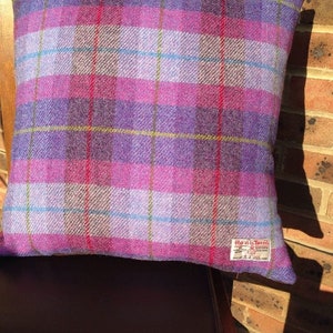May include: A square decorative pillow with a plaid pattern in shades of purple, pink, and blue. The pillow is made of a textured fabric and has a tag that reads "Harris Tweed". The pillow is resting on a wooden chair.