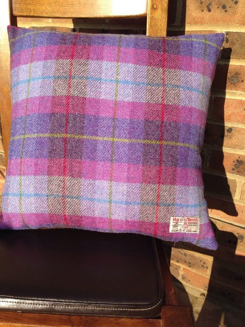 May include: A square decorative pillow with a plaid pattern in shades of purple, pink, and blue. The pillow is made of a textured fabric and has a tag that reads "Harris Tweed". The pillow is resting on a wooden chair.