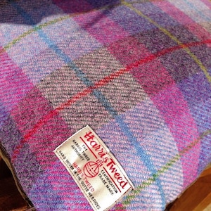 May include: A square cushion with a Harris Tweed label. The cushion features a plaid pattern in shades of purple, pink, blue, and green. The fabric has a textured appearance.