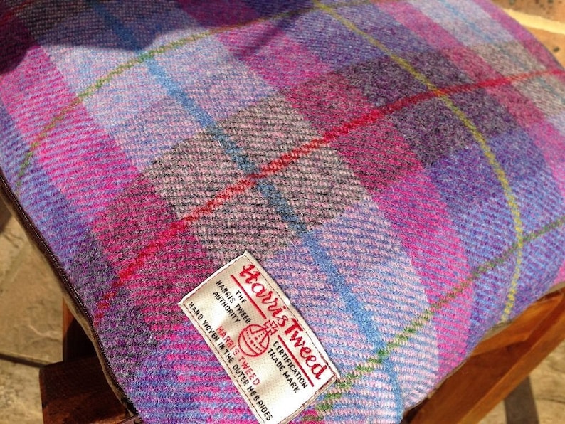 May include: A square cushion with a Harris Tweed label. The cushion features a plaid pattern in shades of purple, pink, blue, and green. The fabric has a textured appearance.