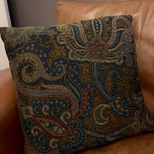 Puede incluir: Una almohada decorativa con un patr&oacute;n de paisley en tonos de azul, marr&oacute;n, dorado y rojo. La almohada est&aacute; sobre un sof&aacute; de cuero marr&oacute;n.