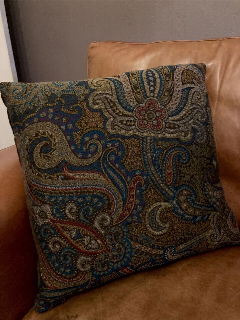Puede incluir: Una almohada decorativa con un patr&oacute;n de paisley en tonos de azul, marr&oacute;n, dorado y rojo. La almohada est&aacute; sobre un sof&aacute; de cuero marr&oacute;n.