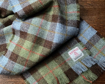 May include: A green, brown, and blue plaid scarf with fringed edges. The scarf has a label that reads "Harris Tweed" with a small logo of a spinning wheel.