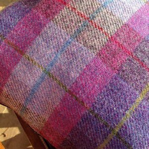 May include: Close-up of a cushion with a vibrant plaid pattern. The fabric features a mix of pink, purple, blue, and yellow lines, creating a colorful and textured design. The cushion is likely made of a woven material, possibly wool or a similar fabric.