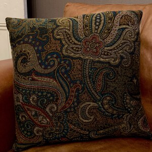 Puede incluir: Una almohada decorativa con un patr&oacute;n de paisley en tonos marrones, azul verdoso y rojo. La almohada est&aacute; hecha de una tela suave y texturizada.