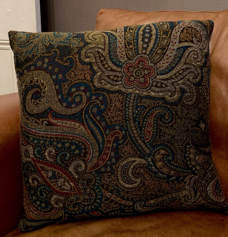 Puede incluir: Una almohada decorativa con un patr&oacute;n de paisley en tonos marrones, azul verdoso y rojo. La almohada est&aacute; hecha de una tela suave y texturizada.
