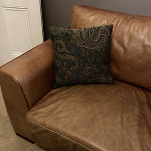 Puede incluir: Un sof&aacute; de cuero marr&oacute;n con un coj&iacute;n de estampado paisley. El sof&aacute; tiene un respaldo capiton&eacute; y un brazo enrollado.