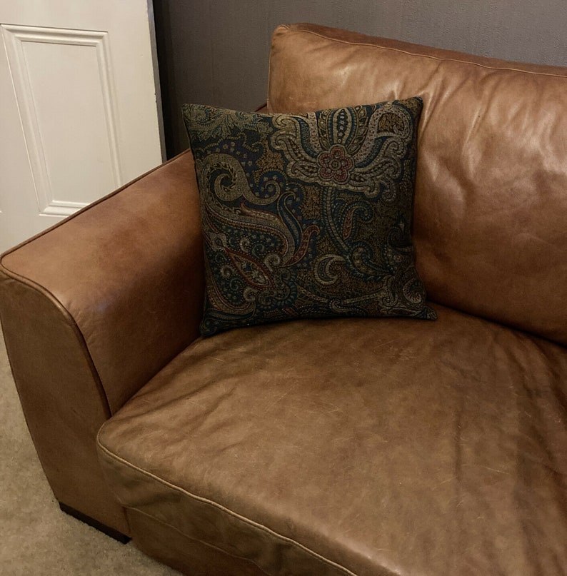 Puede incluir: Un sof&aacute; de cuero marr&oacute;n con un coj&iacute;n de estampado paisley. El sof&aacute; tiene un respaldo capiton&eacute; y un brazo enrollado.