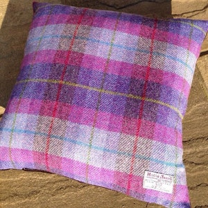 May include: Square decorative pillow with a plaid pattern in shades of purple, pink, and white. The pillow features a Harris Tweed label in the lower right corner. The plaid design includes thin lines of blue, red, and yellow.