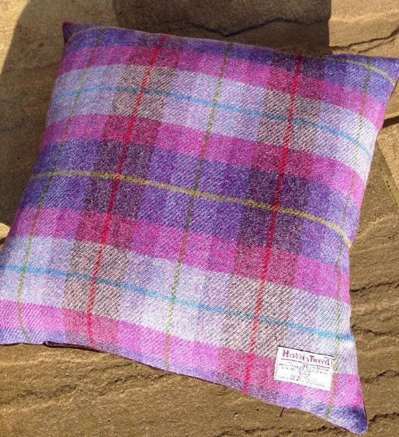 May include: Square decorative pillow with a plaid pattern in shades of purple, pink, and white. The pillow features a Harris Tweed label in the lower right corner. The plaid design includes thin lines of blue, red, and yellow.