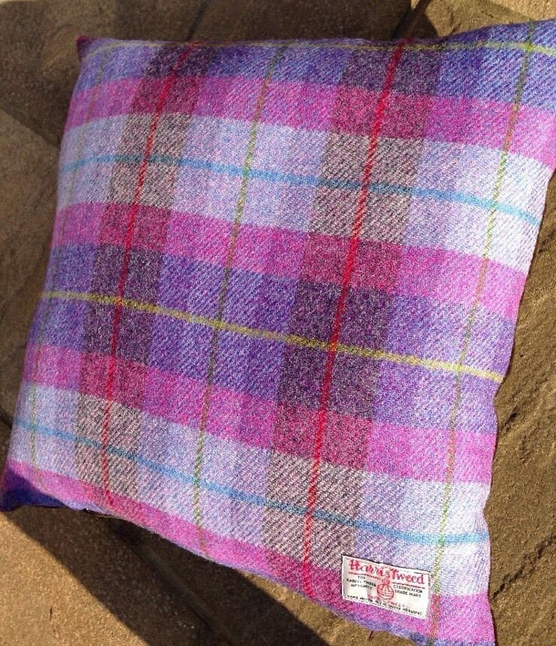 May include: Square pillow with a purple, pink, and white plaid pattern. The pillow has a Harris Tweed label in the lower right corner. The plaid design includes red, yellow, and blue accent lines.