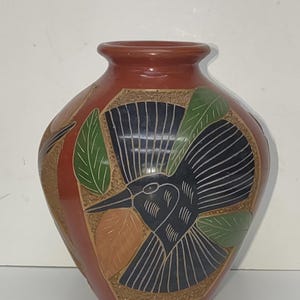 May include: A terracotta-colored ceramic vase with a detailed bird and leaf design. The bird is black with white etched lines, and the leaves are green and orange. The vase has a rounded body and a narrow neck.
