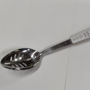 May include: A slotted spoon with a stainless steel bowl and handle. The handle is white with a floral design in gold and green. The spoon is designed for draining liquids from food.