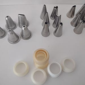 May include: A collection of stainless steel piping tips in various shapes and sizes, including star, round, and leaf tips. Also included are several plastic couplers and rings, all arranged on a white surface. These tools are for cake decorating.