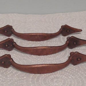 May include: Three antique copper-colored drawer pulls. Each pull has a curved handle with decorative pointed ends and a small circular detail. The pulls are arranged horizontally on a white surface.