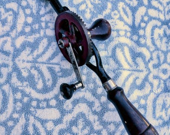 May include: A vintage hand drill with a dark wooden handle and a red gear. The drill has a metal chuck and a crank handle. The tool is set against a blue and white patterned background.