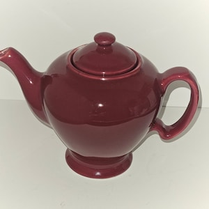 May include: A burgundy ceramic teapot with a lid. The teapot has a rounded body and a spout on one side. The handle is on the opposite side of the spout.