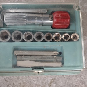 May include: A vintage tool kit in a clear plastic case with a light blue base. The kit includes a silver ratchet handle with a red grip, various sockets, and screwdriver bits. The case is closed with a clear plastic lid.
